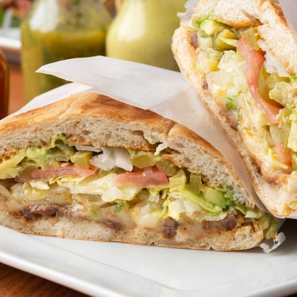 Two halves of a large Mexican torta sandwich filled with meat, lettuce, avocado, and tomato.