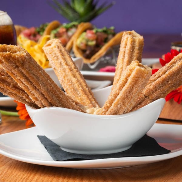 Close-up of cinnamon sugar churros in a white bowl, with tacos and a drink blurred in the background.
