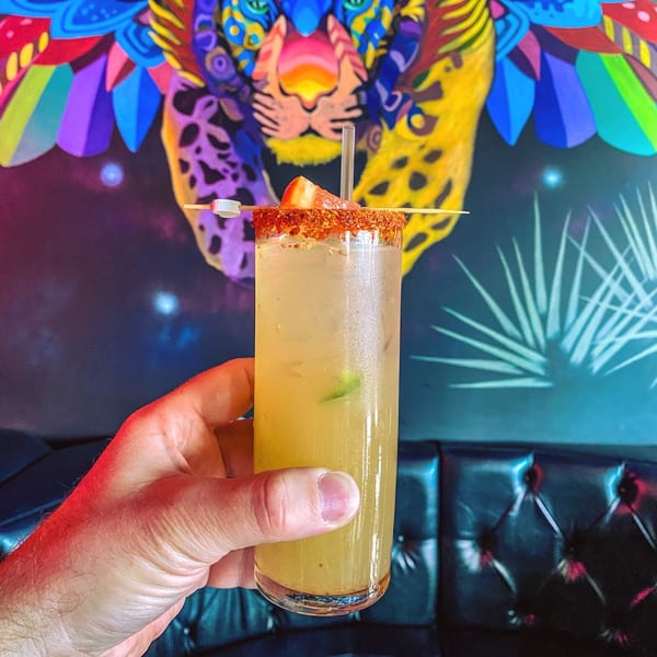 Hand holding a Mezcal cocktail with chili-lime rim and strawberry garnish against a colorful jaguar mural.